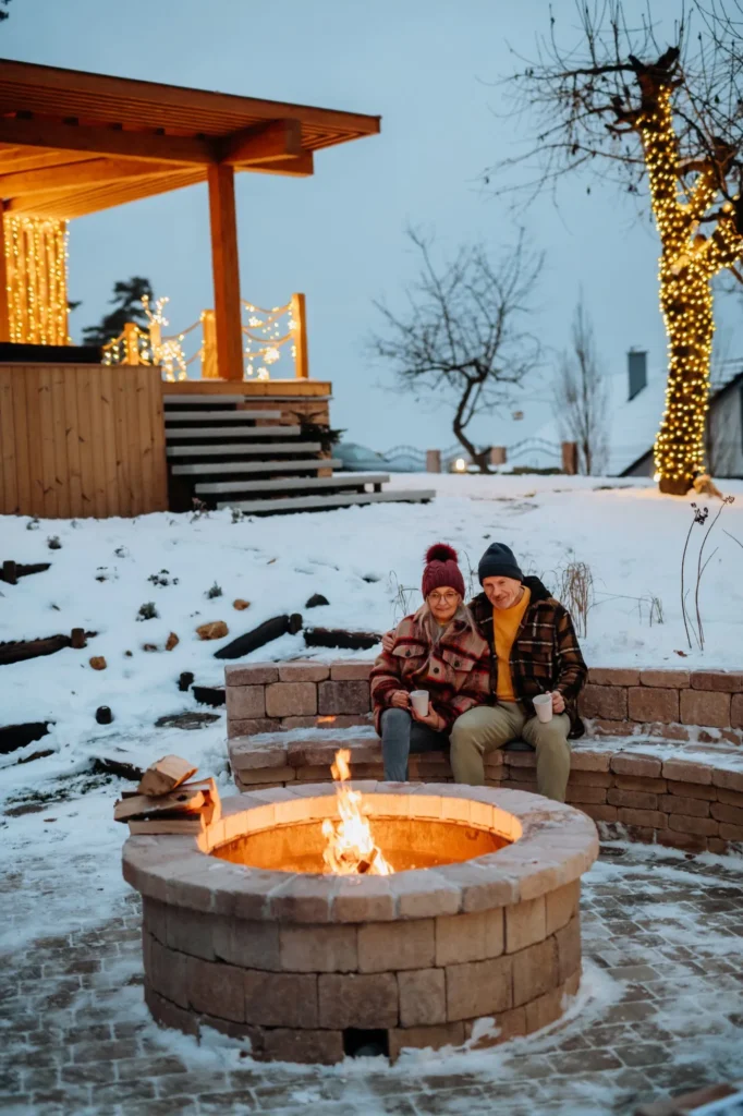 Couple sitting by a stone fire pit in a snowy yard with string lights.